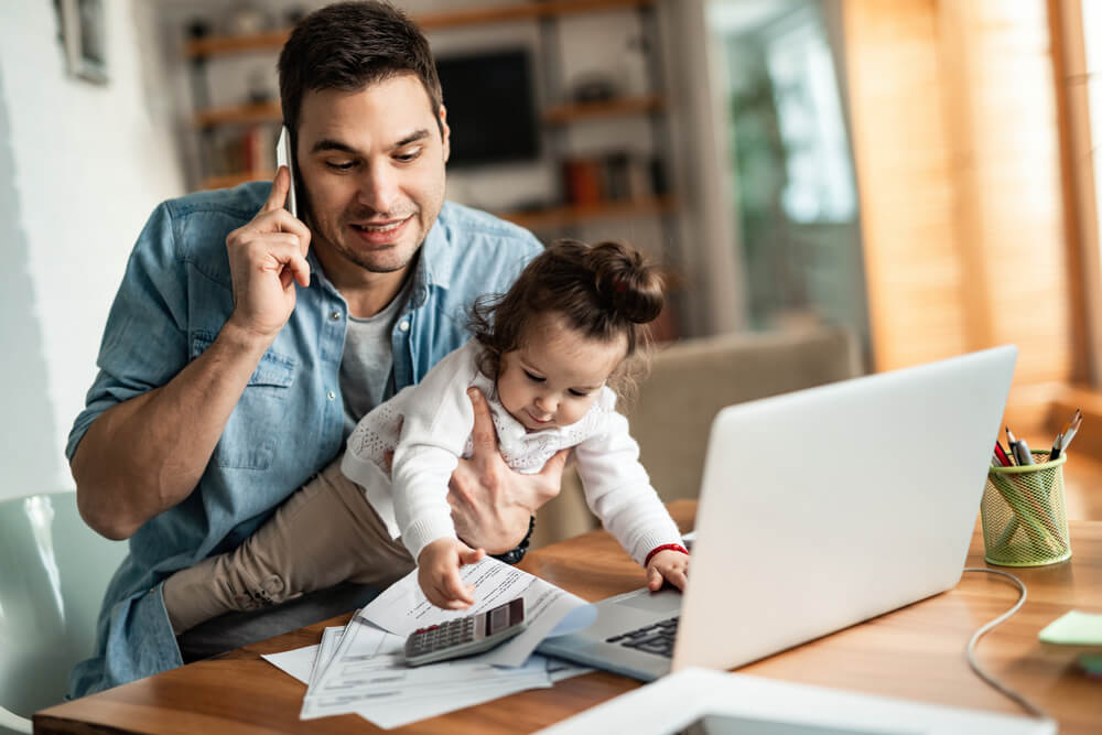 Father working while caring for a toddler. Mile High Psychology Photograph of a father working from home on his laptop, while talking on the phone and holding his daughter, providing example of modern day stresses.